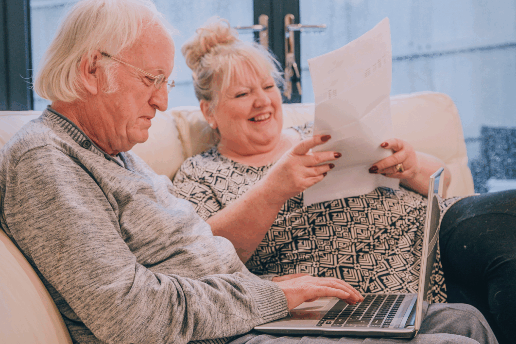 Older couple sitting on the sofa. They are going through bills and working on their laptop. they're smiling