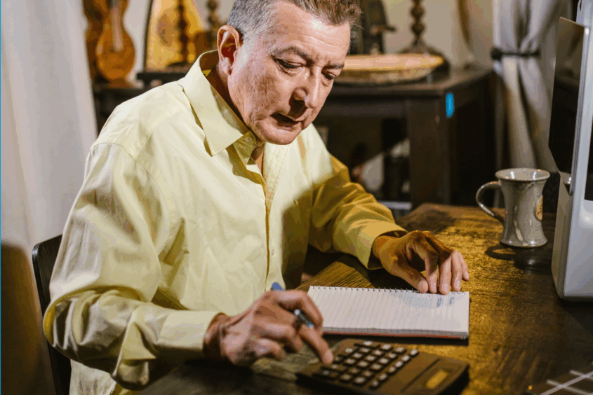 A man sitting at a desk. He has a notepad in front of him and is using a calculator