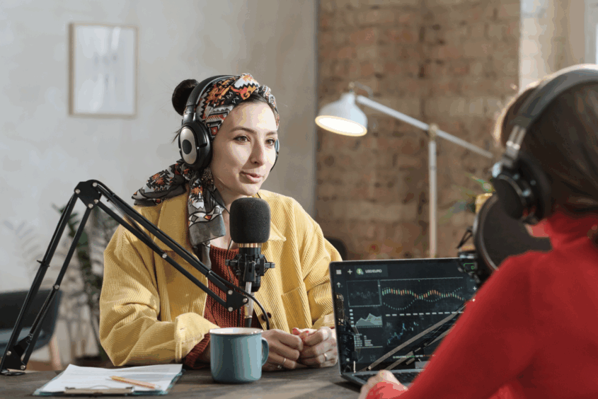 Two women are sat opposite each other across a table. They're recording a podcast. One of the women is wearing a headscarf and is talking into a microphone. The other woman has a laptop with editing software open.