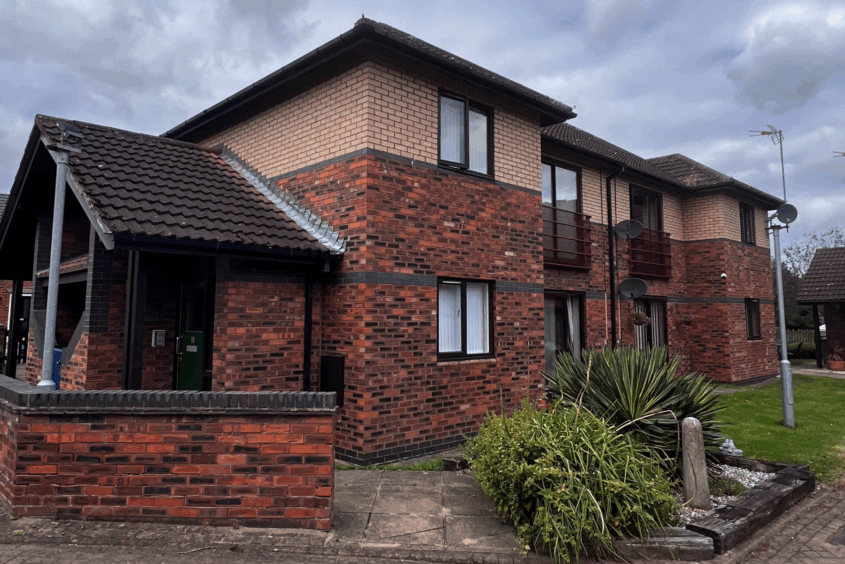 Low-rise residential block in red brick. In front of the building is a lawned area next to shrubbery.