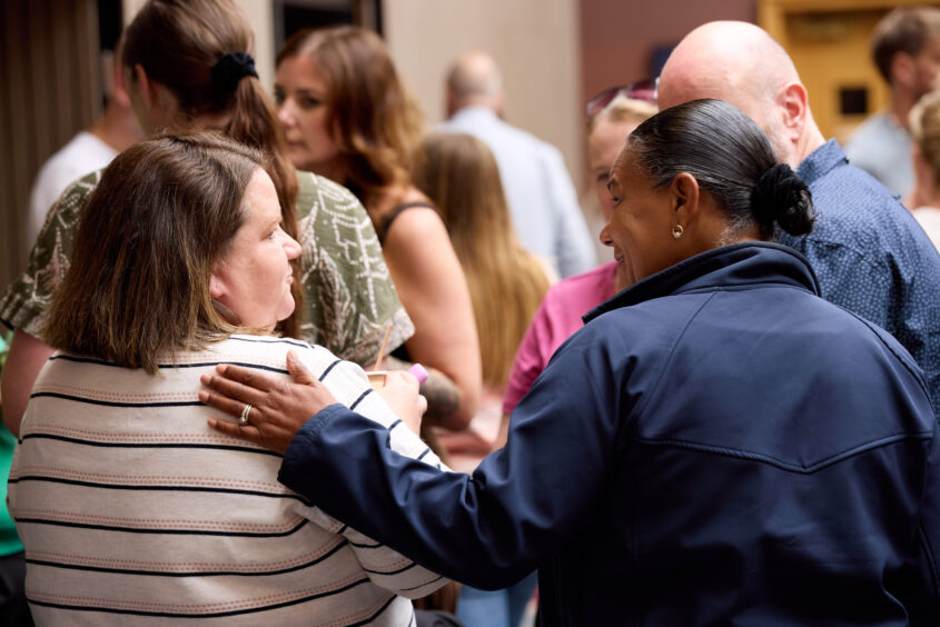 Two women are stood facing away from the camera. One woman has her hand on the shoulder of the other woman in a supportive way. The image supports the 'benefits of working for us' webpage.