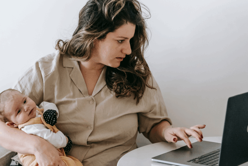 Woman sat at a desk with a laptop in front of her. She is typing on the laptop with one hand. She is holding a baby with her other arm. She is looking stressed.
