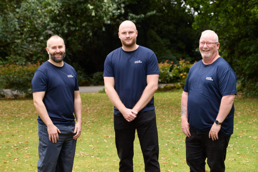 Mike, Charlie and Tony standing on a lawn outside. They are smiling. There are shrubs and trees in the background.
