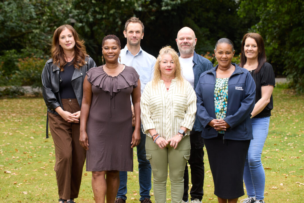 North team standing on a lawn together. They are smiling. There are trees in the background.
