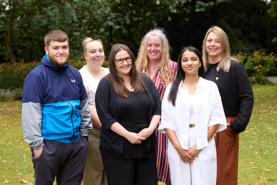Customer experience team standing on a lawn together. They are smiling. There are trees in the background.