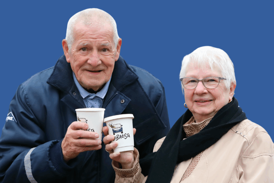 An older couple are outside weating winter clothes. They are each holding up a cup of coffee as if to say 'cheers'. They are both smiling.