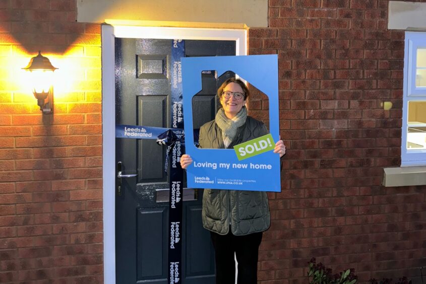 A customer is standing outside the front door of her new home. She is smiling and holding up a selfie board with a home shape cut out. The selfie board says 'loving my new home'