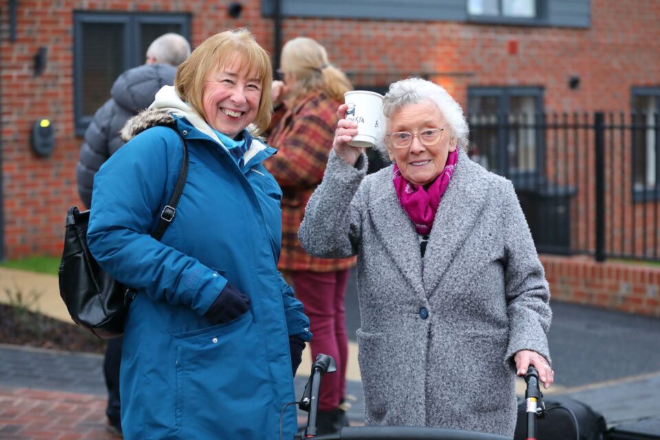 Two women stood next to each other. One woman is holding a cup of coffee up saying cheers.