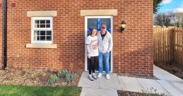 A couple standing outside the front door of their new home. They have an arm around each other and are smiling.