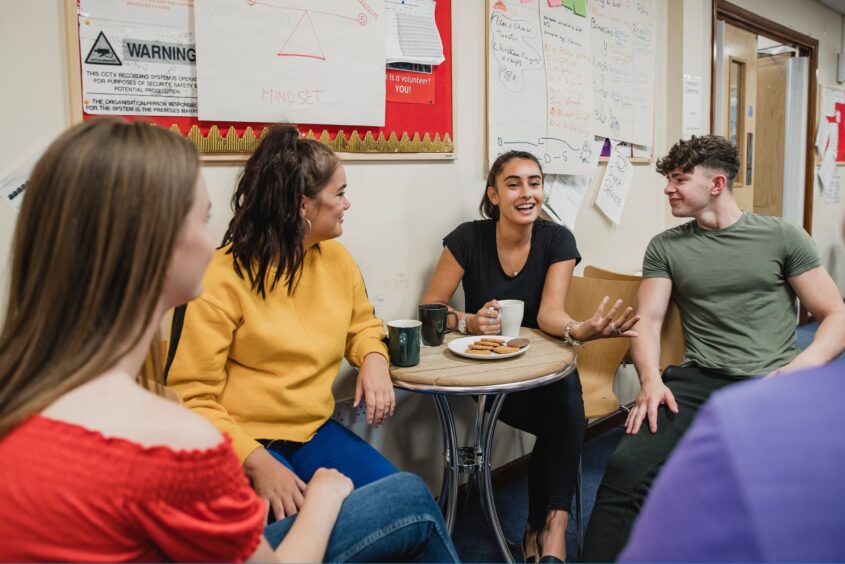Four young people sat around a table in a cafe. They are chatting, drinking coffee and eating biscuits.