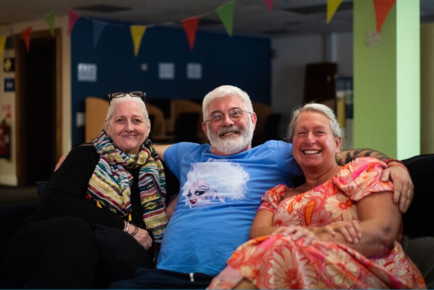 Three people are sat together on a sofa in a community hall. The man in the middle has his arms around the people on either side of him. There's bunting in the background and everyone is smiling