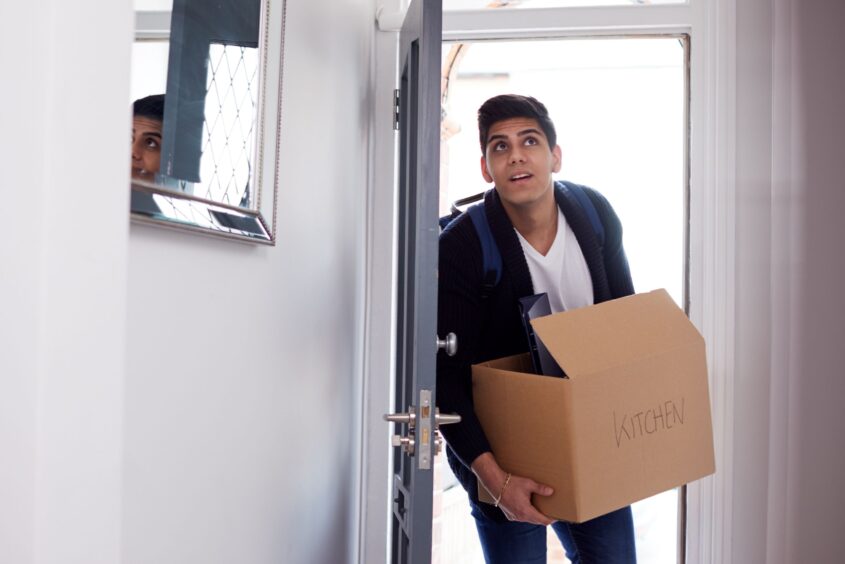 Young person walking through an open front door of their new home. They are carrying a cardboard box that has 'kitchen' written on it.
