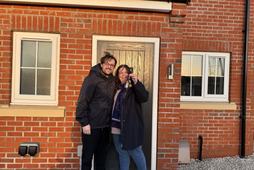 A couple are standing outside the front door of a new build red bring home. They are holding their keys up and smiling