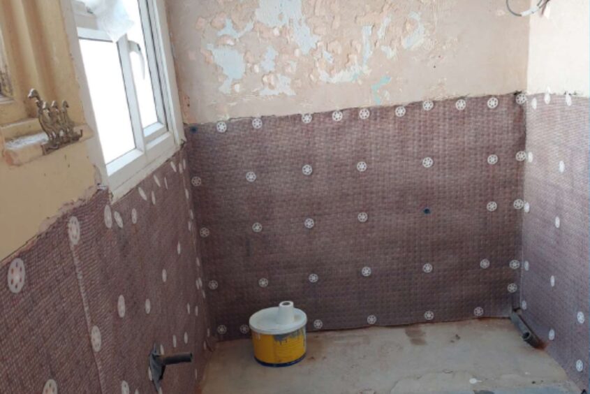 Three walls of the bathroom. The top half of the walls has bare plaster, the bottom half shows a damp-proof membrane that's just been installed.