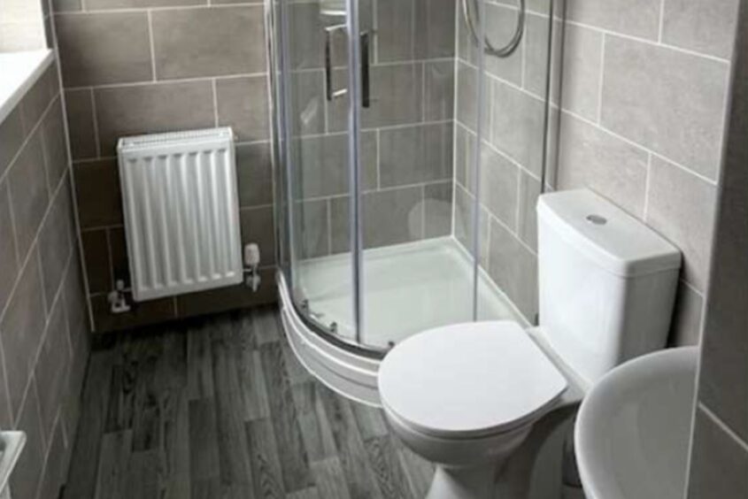 A new bathroom suite in someone's home. There is a toilet shower and sink on the right hand wall and a radiator on the back wall. The walls are tiled and there's lino on the floor.