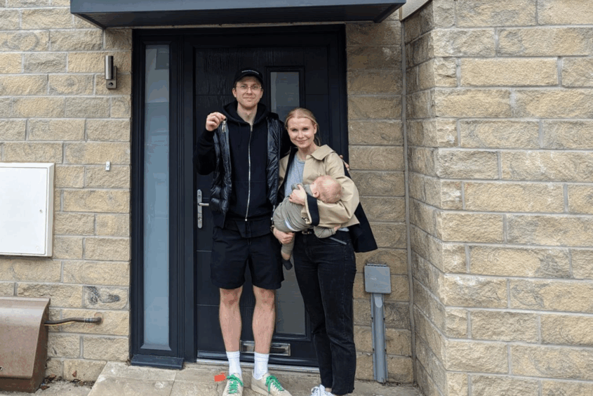 A couple are standing outside the front door of their new home. The woman is holding their baby, The man is holding up the keys to the door. The couple are both smiling.