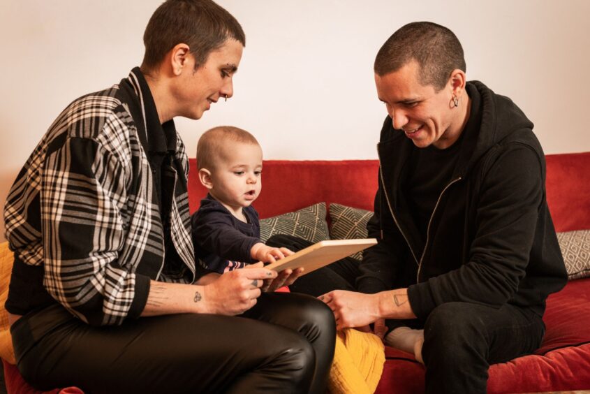 A couple are sitting on the sofa with their toddler. They are playing with an electronic tablet.