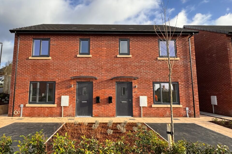A red brick semi-detached new build home. There's a car parking space and shrub area in front of each home