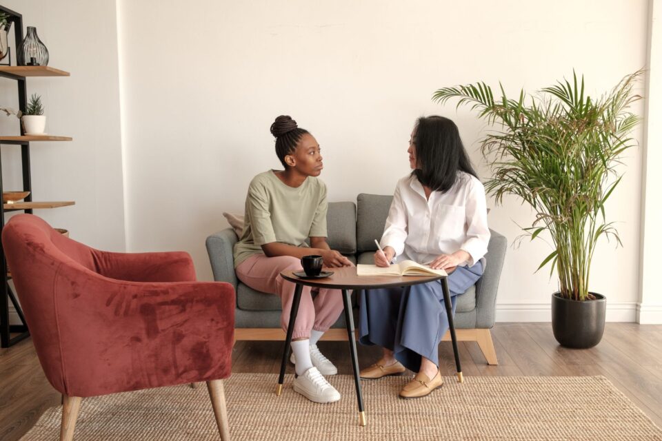 Two people sat on a sofa. The woman is taking notes of what the other woman is saying.