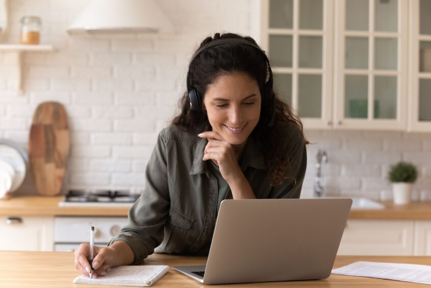 Woman sitting in her kitchen. She is using a laptop, making notes and wearing a headset. She is smiling.
