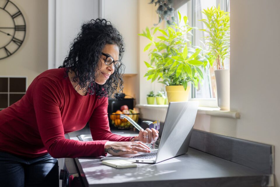 A woman leaning on the kitchen worktop looking at her laptop with bills around her