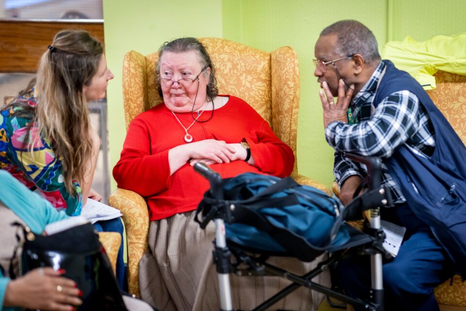 Three people are sitting together in a community building. They are all chatting. One person is leaning on a walking support.