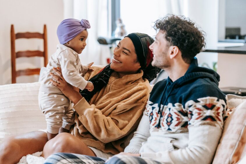 A man and woman are sitting on the sofa. The man is wearing a winter jumper and the woman is holding a baby.
