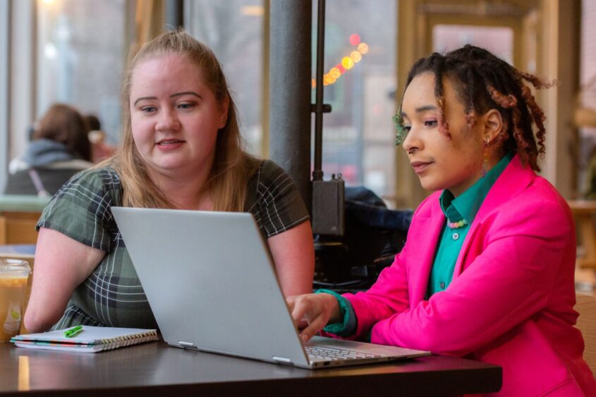 Two people sit together at a cozy café table, collaborating on a laptop. The woman on the left, wearing a green plaid dress, has a disability and her lower arms are missing. She is engaged in the conversation, with a notebook, pen, and drink nearby. The person on the right, dressed in a bright pink blazer over a green shirt, gestures toward the laptop screen. The background is softly lit, with blurred figures suggesting a warm, bustling atmosphere.