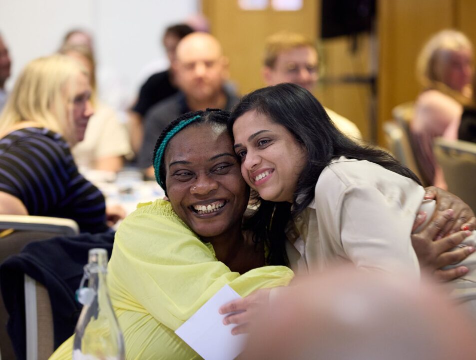 Two colleagues hugging and smiling. In the background there are blurred images sat around tables as though at a conference