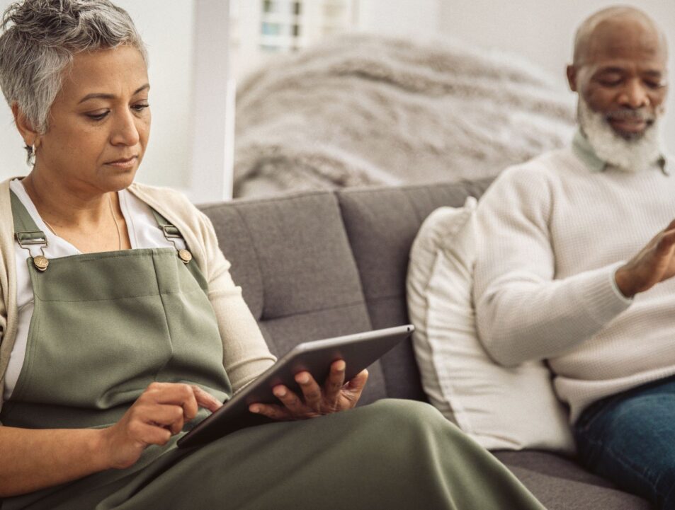 Two people sat on a sofa. One is a woman looking at her ipad and next to her is a man looking at his phone.