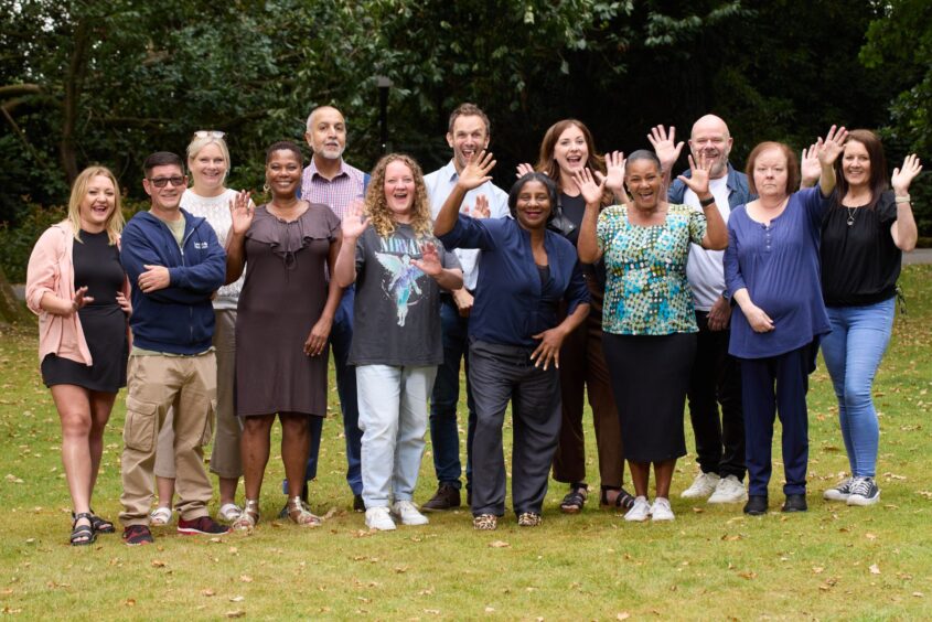 Group of people waving at the camera, stood outside on grass.
