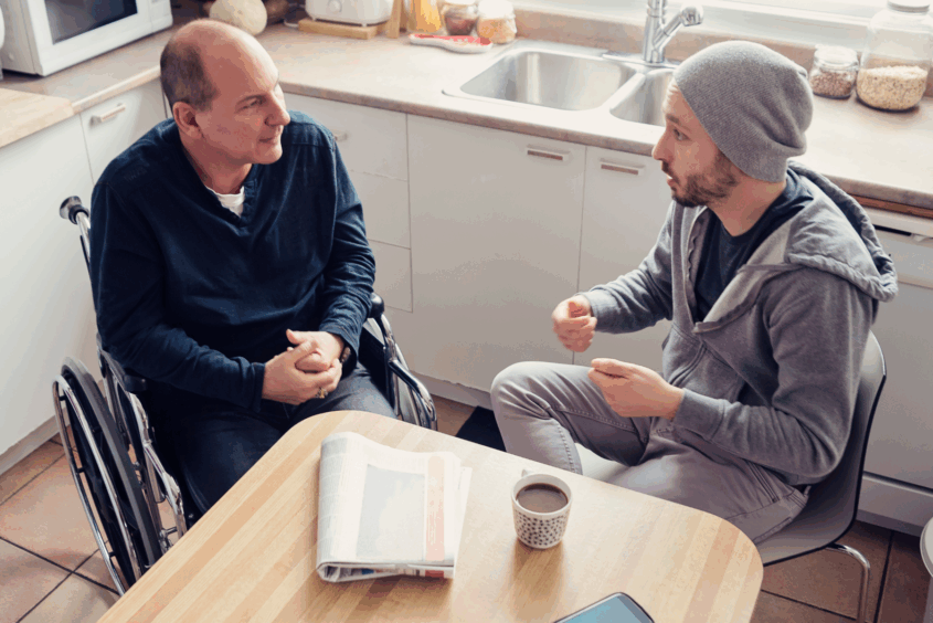 Two men are chatting in a kitchen. One is using a wheelchair, the other is sat on a chair.