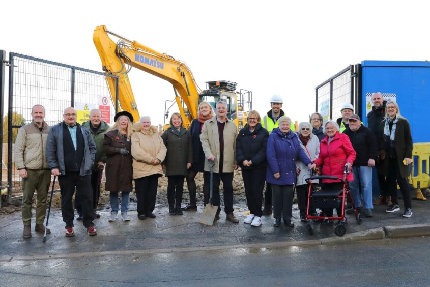 People stood together in a row. They are all wearing coats and smiling at the camera. They are stood in front of a construction site with a digger behind them.