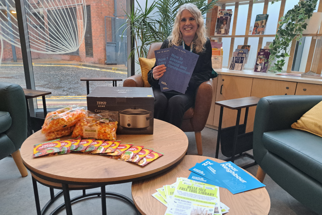 Chairs are arranged around a small table which holds leaflets, stew ingredients and an air fryer. Someone is sat on one of the chairs holding up more leaflets and smiling