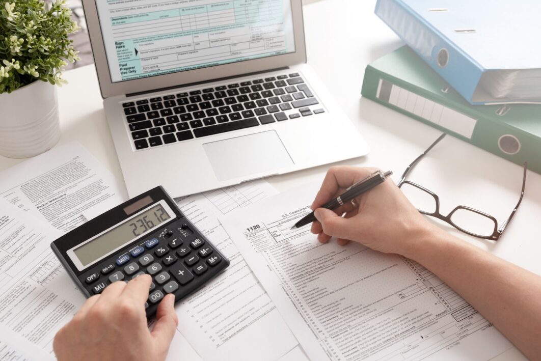 Person working at a laptop using a calculator and looking at paperwork
