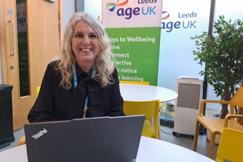 Woman sat behind a laptop at a table, smiling at the camera