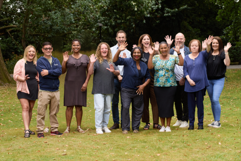Group of people waving at the camera, stood outside on grass.