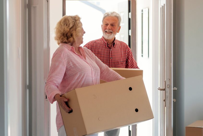 Two older people walking through a door carrying cardboard boxes