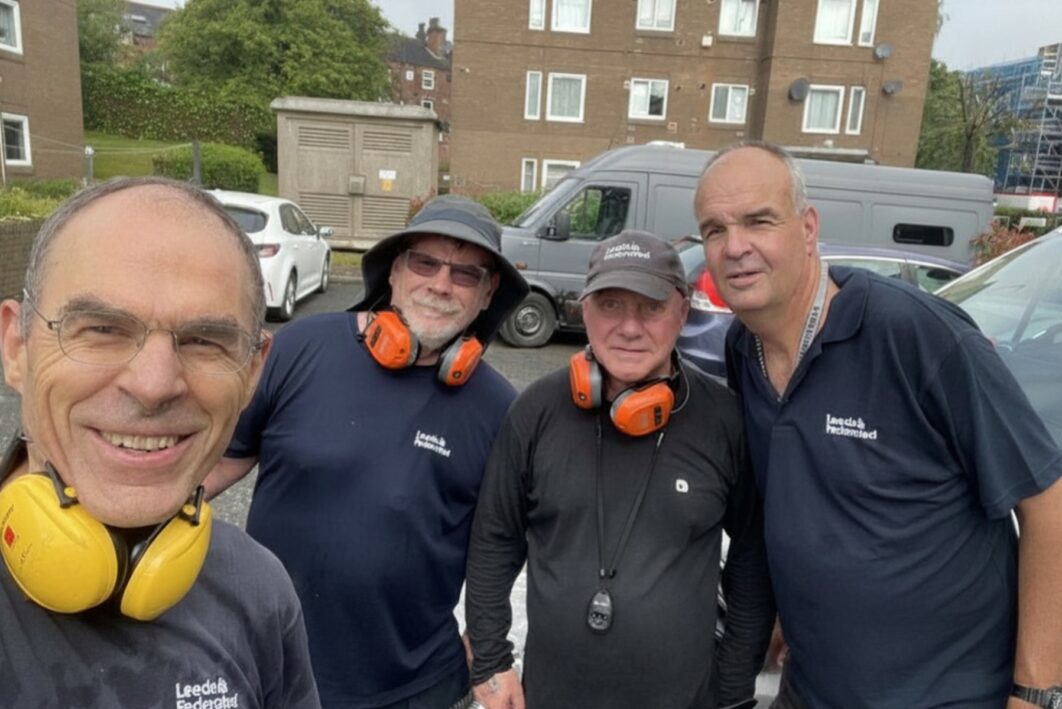 4 men stood together smiling for a photo. They have Leeds Federated t shirts on and ear protection around their necks.