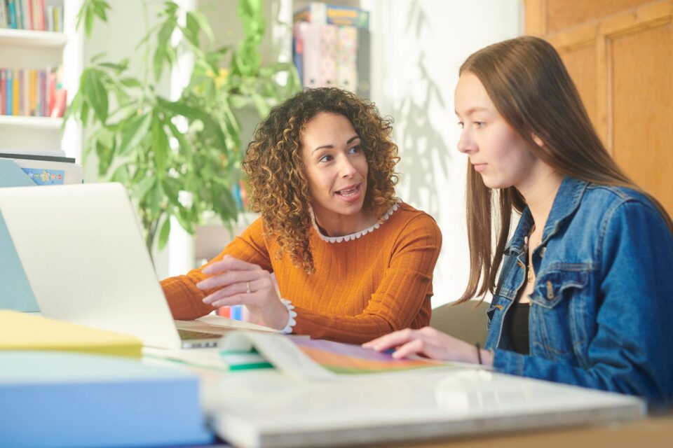 Two women sat next to each other - a woman has a laptop and looks like she is explaining to the other women something